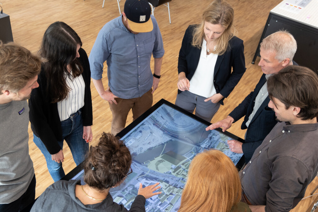 Gesprächssituation mit mehreren Personen rund um einen Touchtisch, auf dem ein digitales Stadtmodell abgebildet ist.